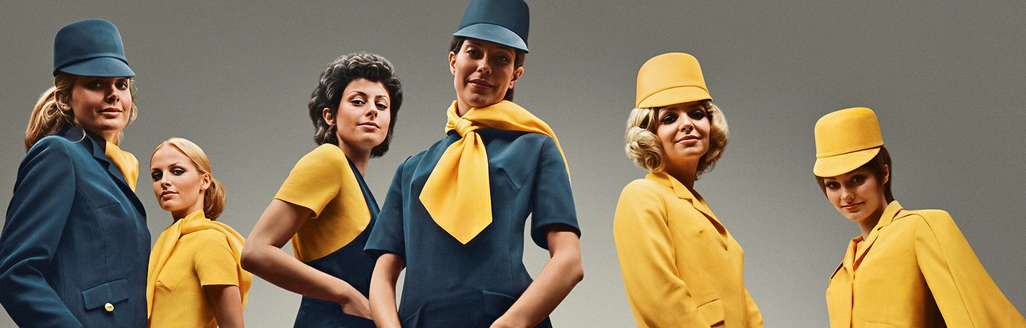 A group of flight attendants wearing vintage blue and yellow uniforms with matching hats.