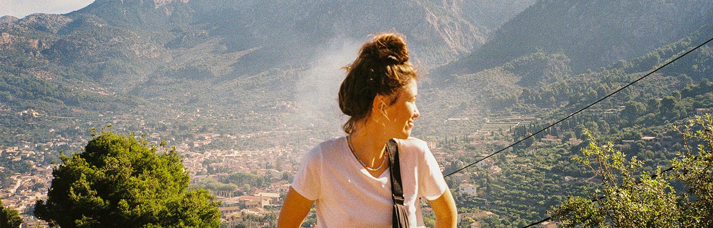 Frau, dreht sich seitwärts weg aus dem Bild, im Hintergrund ist ein typisches mallorquinies Bergdorf zu sehen