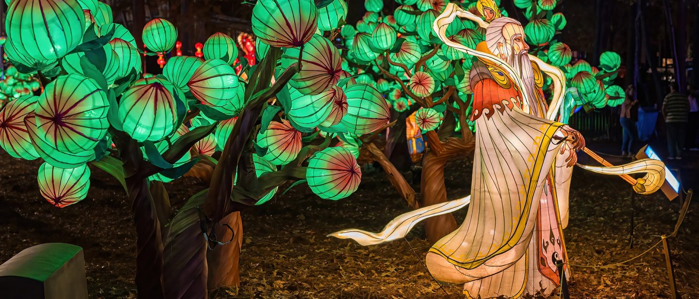 An elaborate lantern figure of an old man with a long beard stands among glowing green, tree-like lanterns.