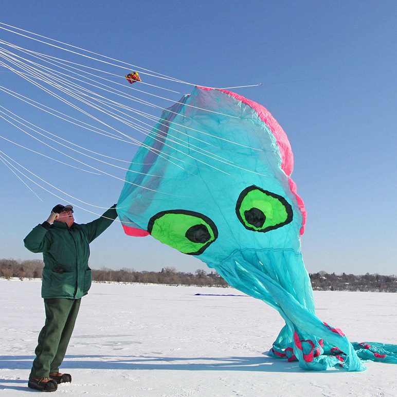 A man on a snowy surface holds a large, turquoise squid-shaped kite with bright green eyes.