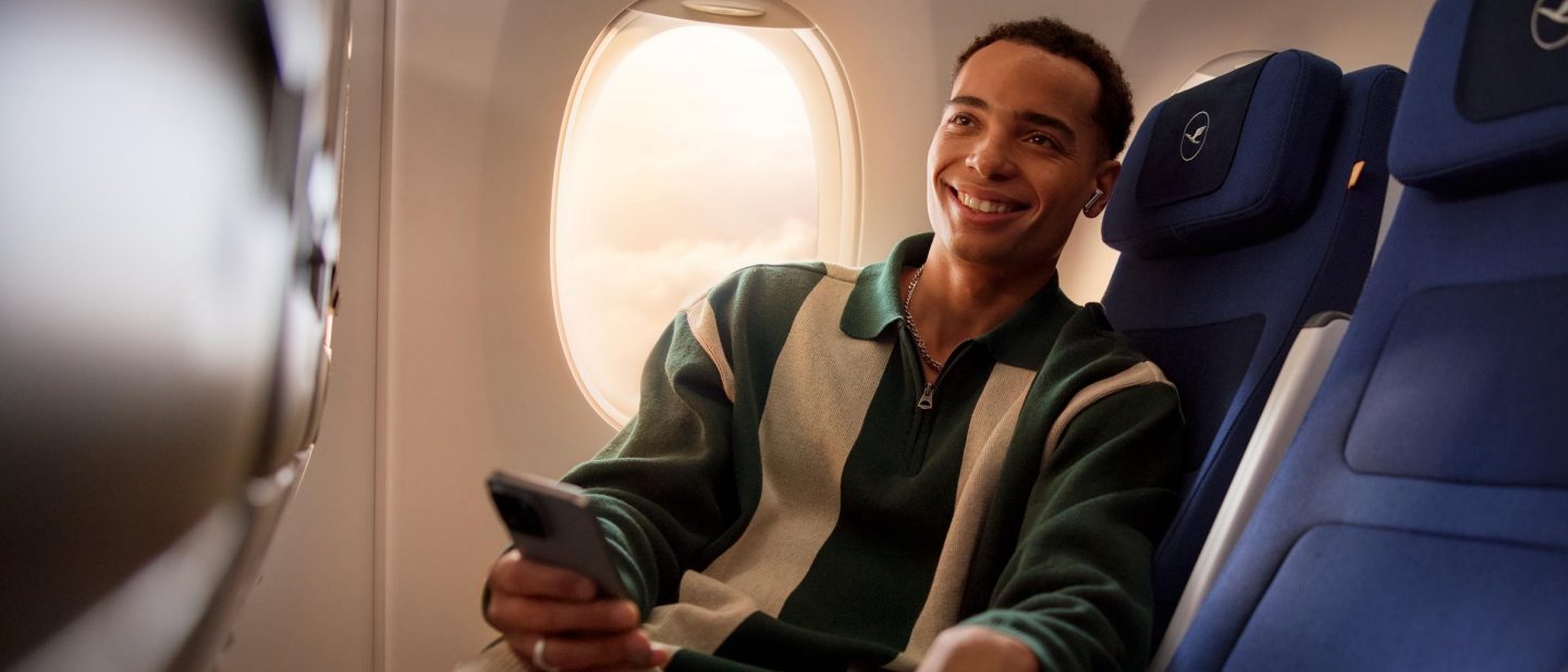 Happy traveler smiles from a Lufthansa window seat, phone in hand.