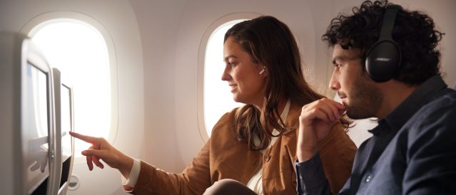 Passenger smiles, holding phone, wearing earbuds by window on a Lufthansa flight.