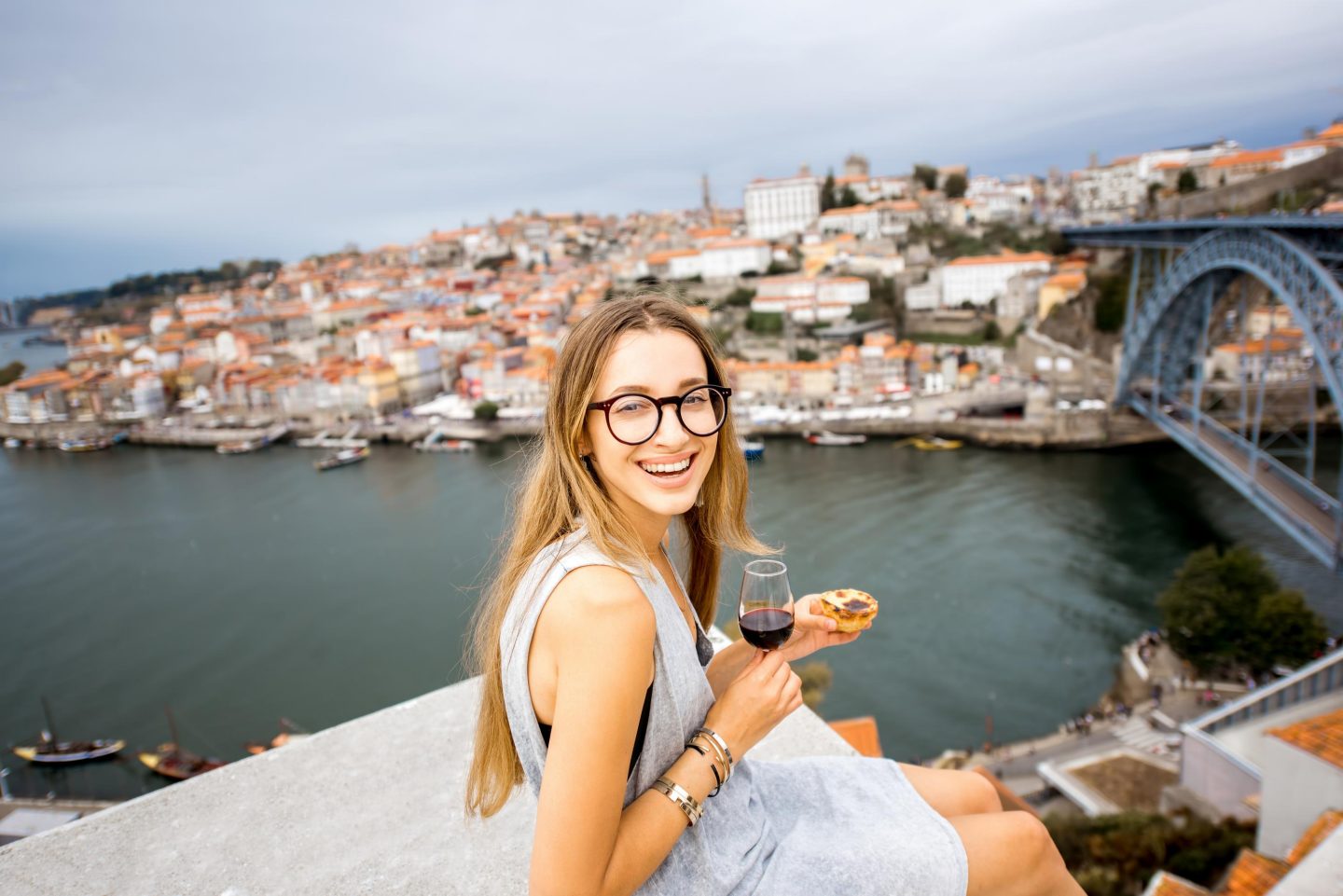 Portugal, Porto: sitzende Frau blickt auf den Fluss und die Skyline w&radic;&sect;hrend sie Rotwein und portugiesischen Pastel de Nata in den H&radic;&sect;nden h&radic;&sect;lt // Portugal, Porto: Seated woman looks out over the river and the skyline while holding red wine and Portuguese Pastel de Nata in her hands