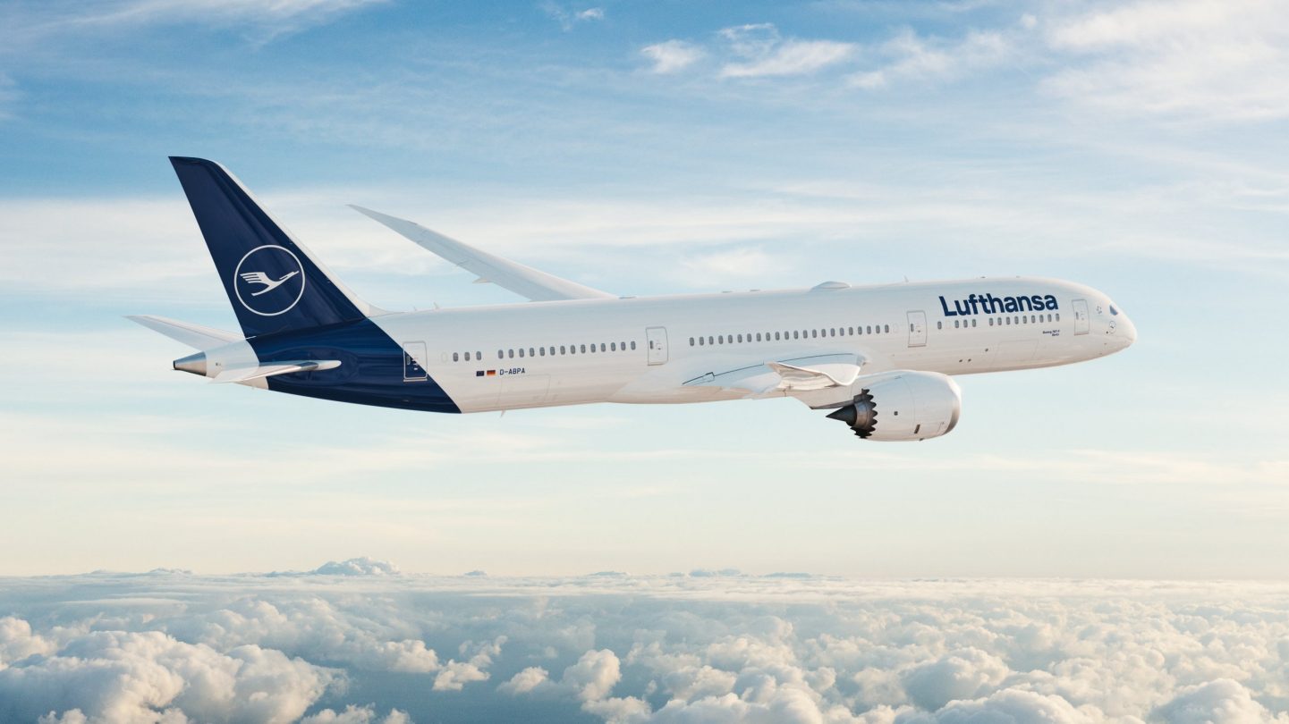 A Lufthansa passenger plane flying above a sea of white clouds in a bright blue sky.