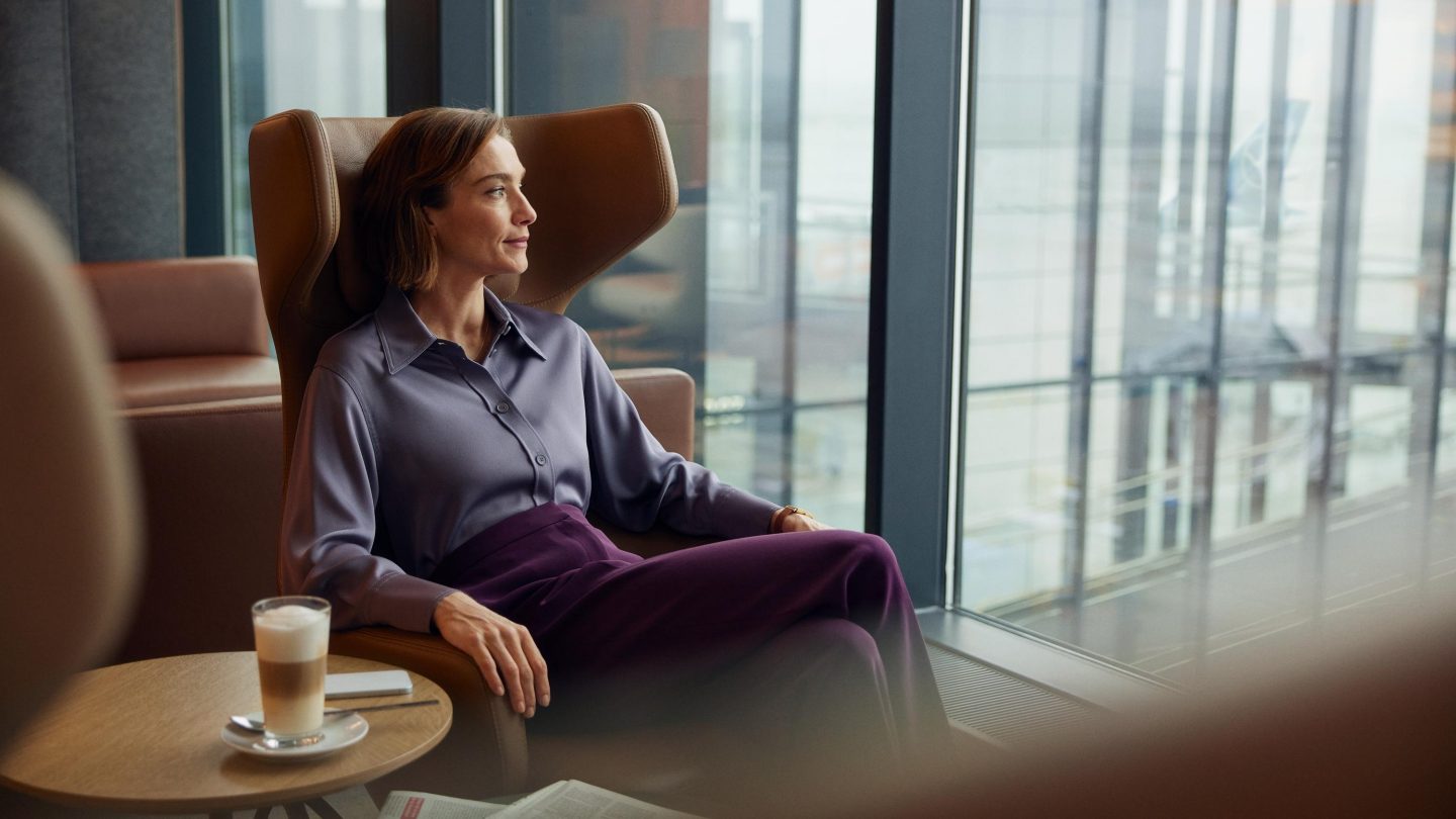Person seated in a modern office lounge chair beside a table with a coffee, looking out through large floor-to-ceiling windows.