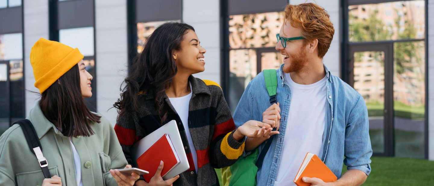 Eine Gruppe l&radic;&sect;chelnder Studenten geht mit B&radic;&ordm;chern und Rucks&radic;&sect;cken auf dem Universit&radic;&sect;ts Campus spazieren und unterh&radic;&sect;lt sich. // Group of smiling students with books and backpacks walking on university campus, talking.