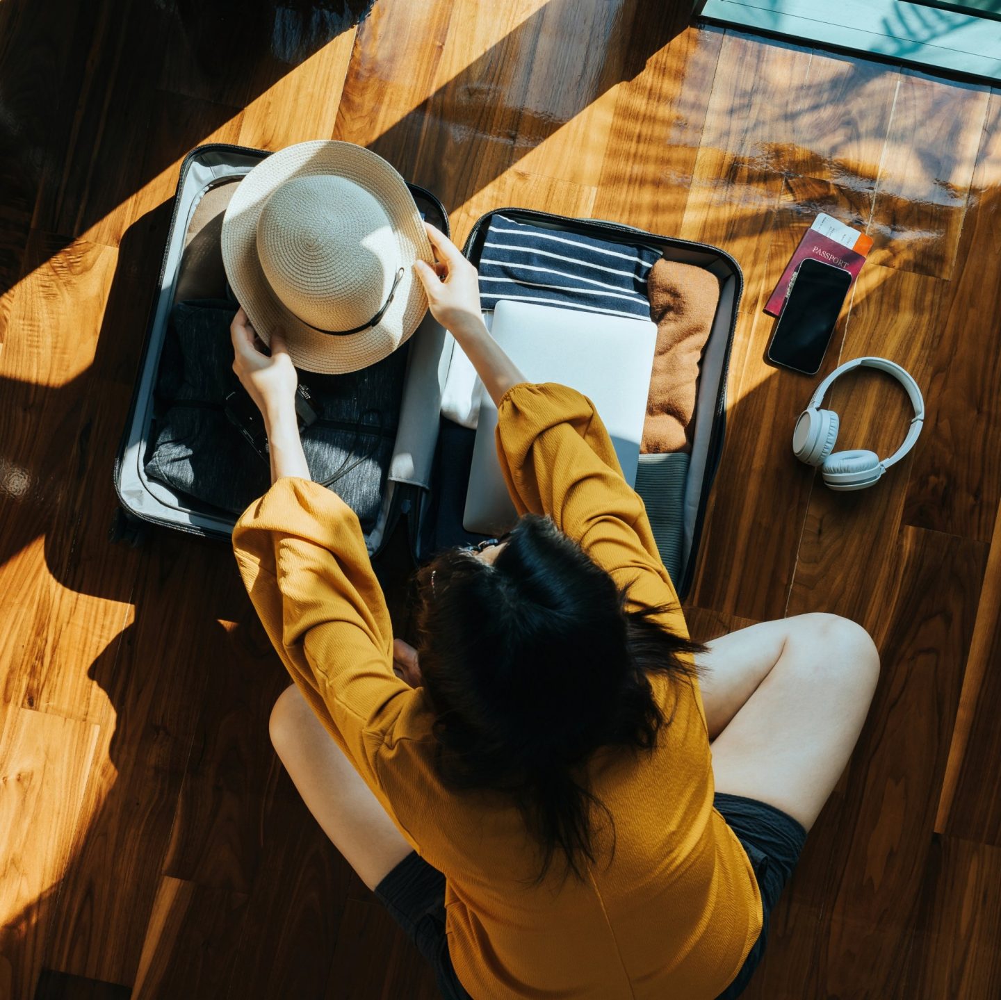 Blick von oben auf eine junge Frau, die in ihrem Schlafzimmer auf dem Boden sitzt und einen Koffer f&radic;&ordm;r eine Reise packt. Sie macht sich f&radic;&ordm;r den Urlaub bereit. // Overhead view of young woman sitting on the floor in her bedroom, packing a suitcase for a trip. Getting ready for a vacation.