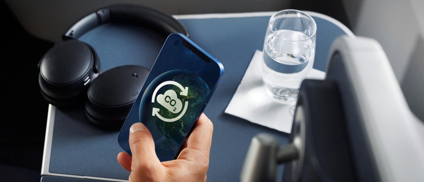 Ein Mann nutzt sein Smartphone und die pers&ouml;nliche Ablage in der Business Class mit Kopfh&ouml;rern, einer Schale mit N&uuml;ssen und ein Wasserglas sind zu sehen. Auf dem Smartphone-Display sehen wir ein CO2 compensation Logo. // A man uses his smartphone and the personal tray in business class with headphones, a bowl of nuts and a glass of water can be seen. On the smartphone-screen we see the CO2 compensation logo.