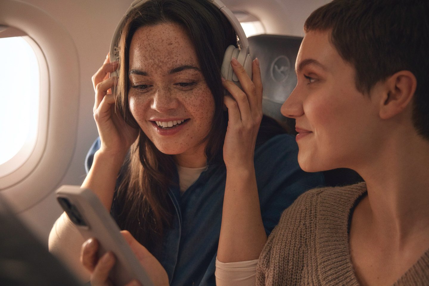 Two smiling women enjoying their flight with headphones and a phone.