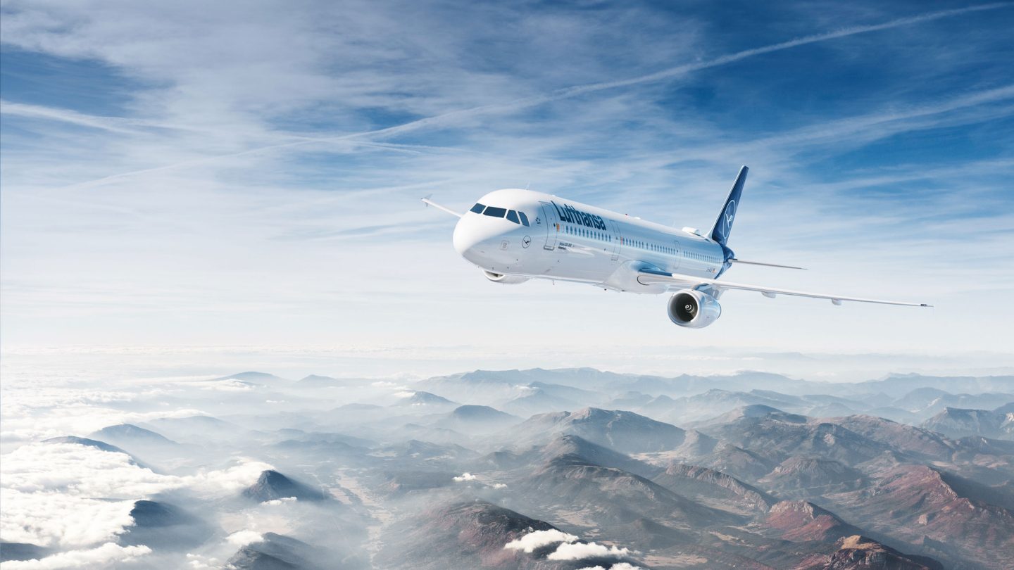 Un avion Lufthansa survole des montagnes brumeuses sous un ciel bleu clair.
