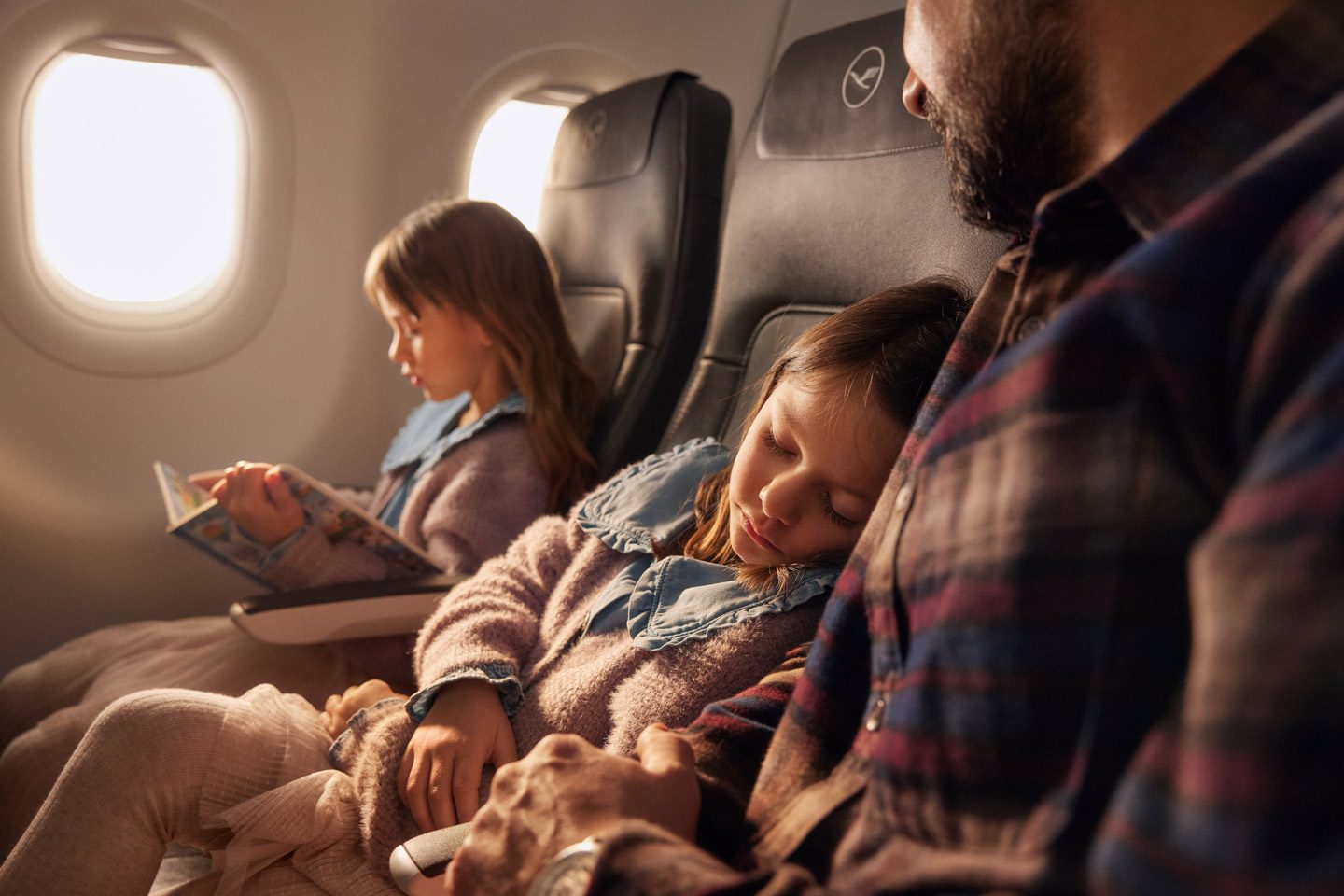 Family comfort on flight: girl asleep on shoulder, another reads by window.