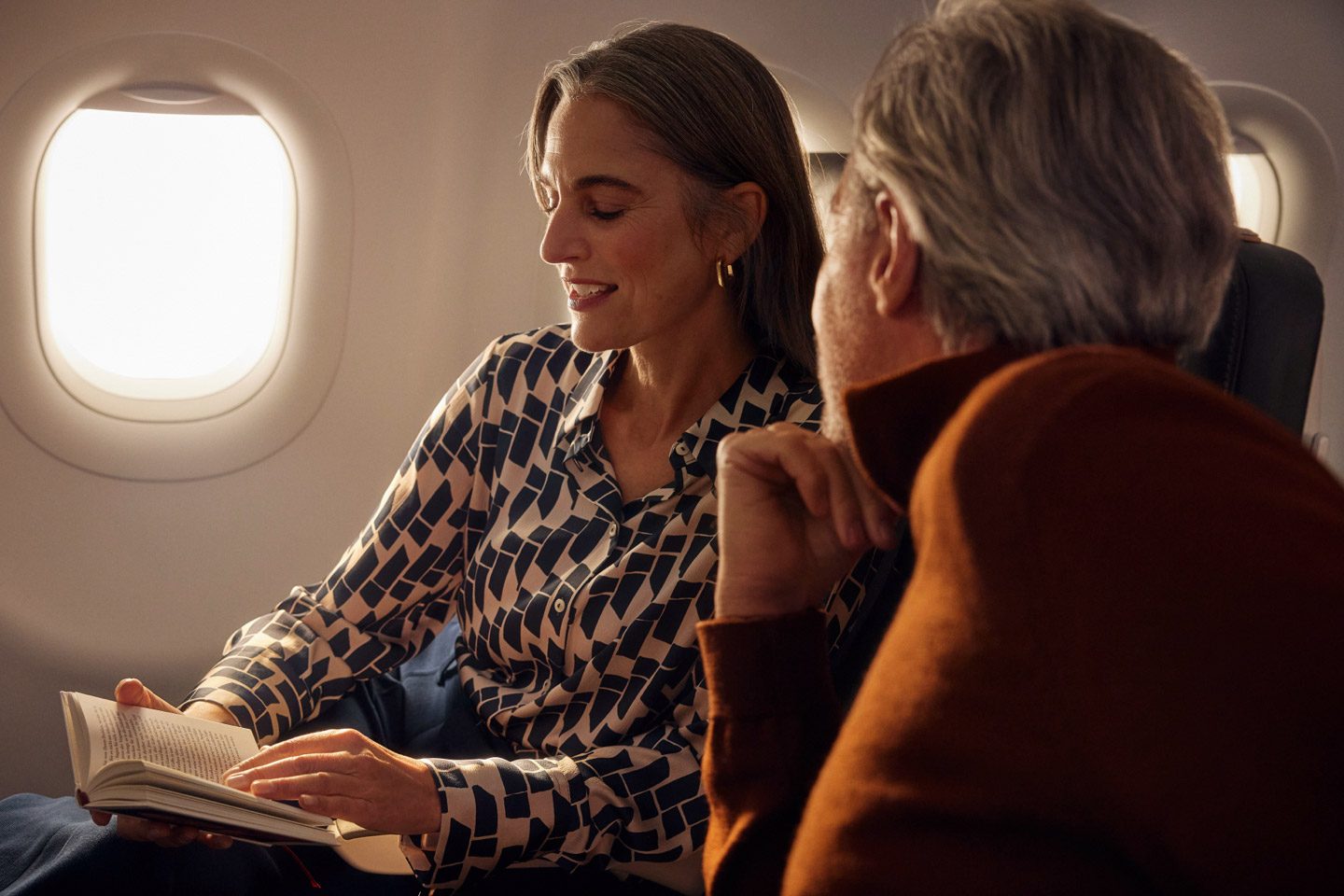 A woman is reading and smiling at the person sitting next to her on the plane.