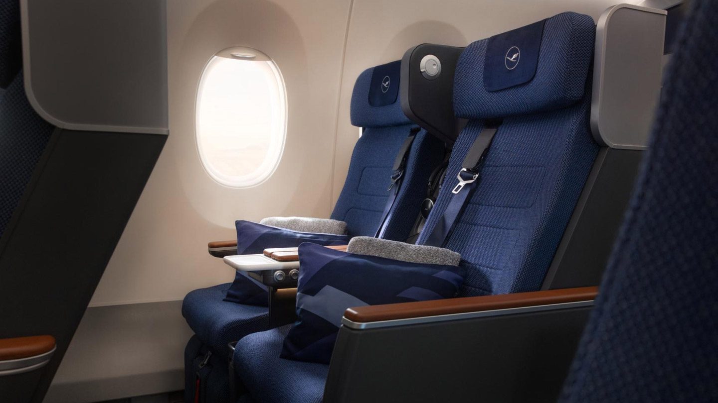 Modern blue airplane seats with Lufthansa headrests, comfort amenities, and a bright window view.
