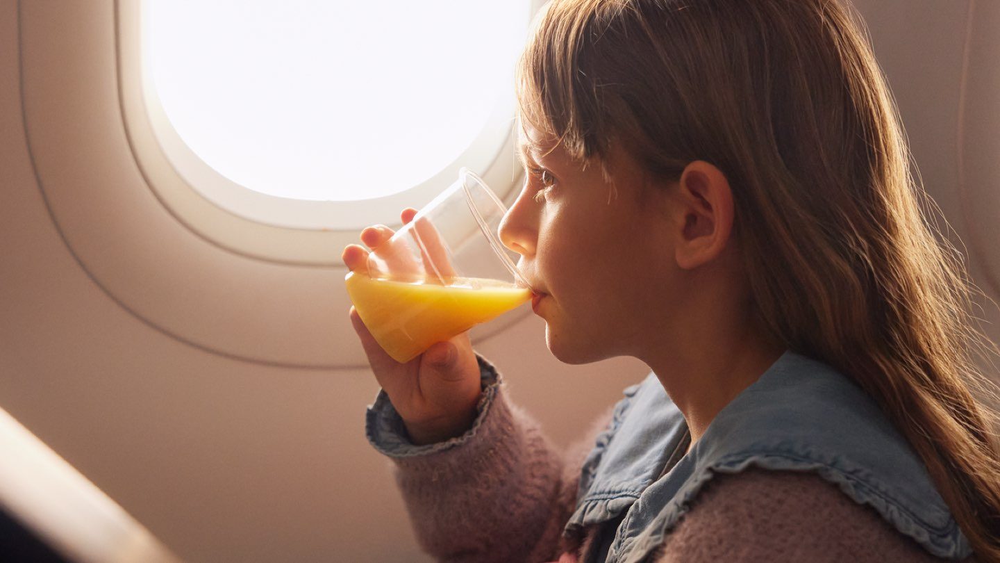 Ein junges Mädchen sitzt am Flugzeugfenster, genießt Orangensaft und liest ein Buch.