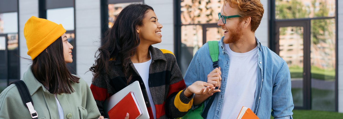 Group of multiracial smiling students with books and backpacks walking on university campus, talking. Emotional friends wearing casual clothing communication together outdoors. Education concept