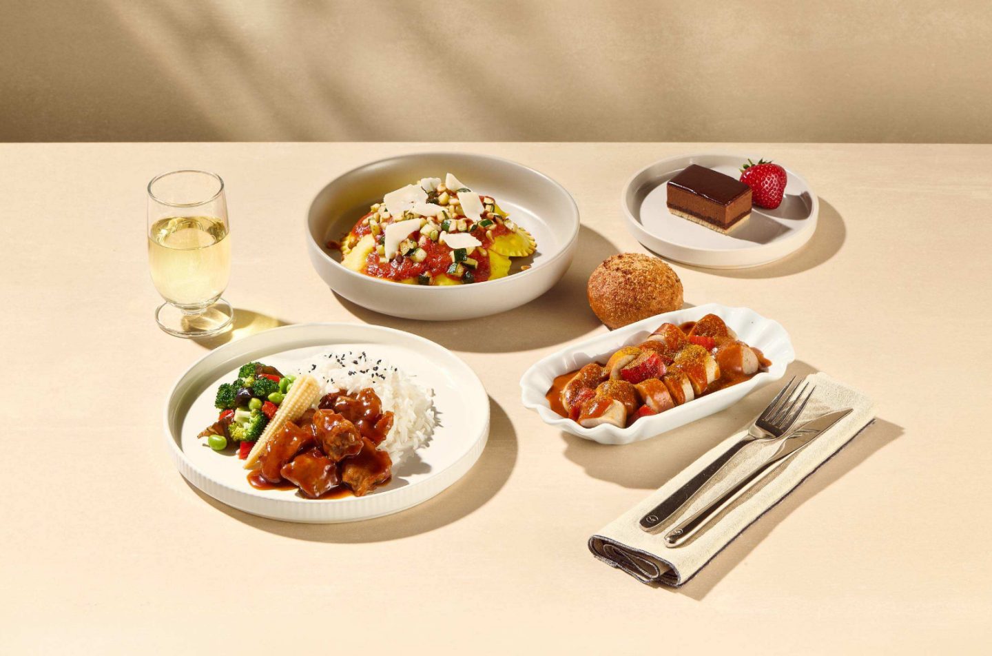 Platos variados, concretamente unos raviolis con salsa de tomate, arroz con verduras y carne, unas rodajas de salchicha con salsa, un panecillo, un postre de chocolate con una fresa y una copa de vino blanco.