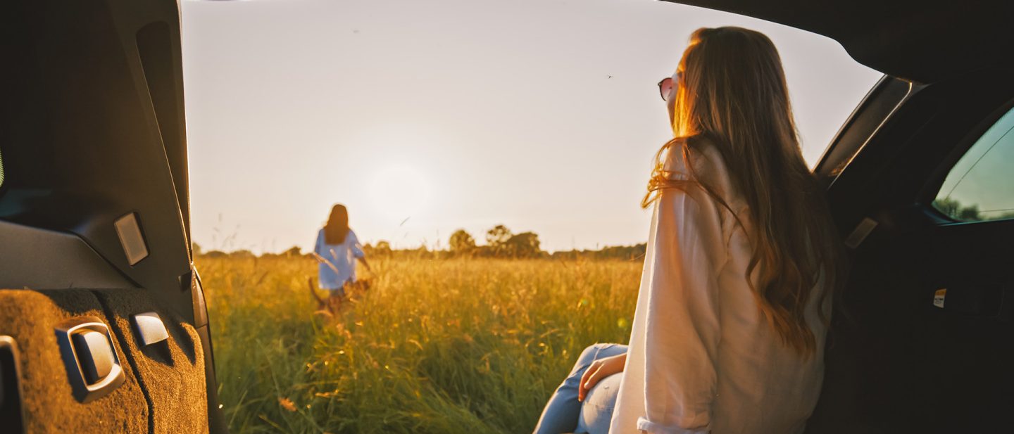 Frau im offenen Kofferraum blickt bei Sonnenuntergang auf ein goldenes Feld mit einer Person in der Ferne.