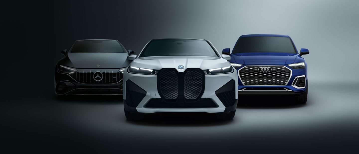 Front view of three cars from different brands against a dark background: a black Mercedes on the left, a white BMW in the center, and a blue Audi on the right.