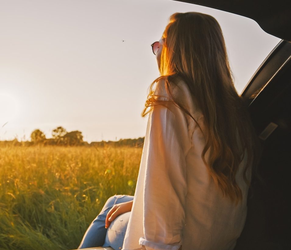 Frau mit langen Haaren sitzt im offenen Kofferraum eines Autos und blickt bei Sonnenuntergang über ein goldenes Grasfeld.