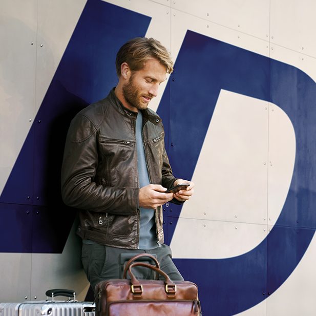 Ein Mann mit einem Koffer blickt auf sein Handy vor einem großen „ID“-Logo.