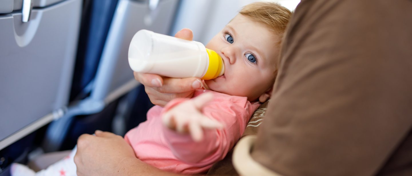 Dad holding his baby daughter during flight on airplane going on vacations. Baby girl drinking formula milk from bottle. Air travel with baby, child and family concept.