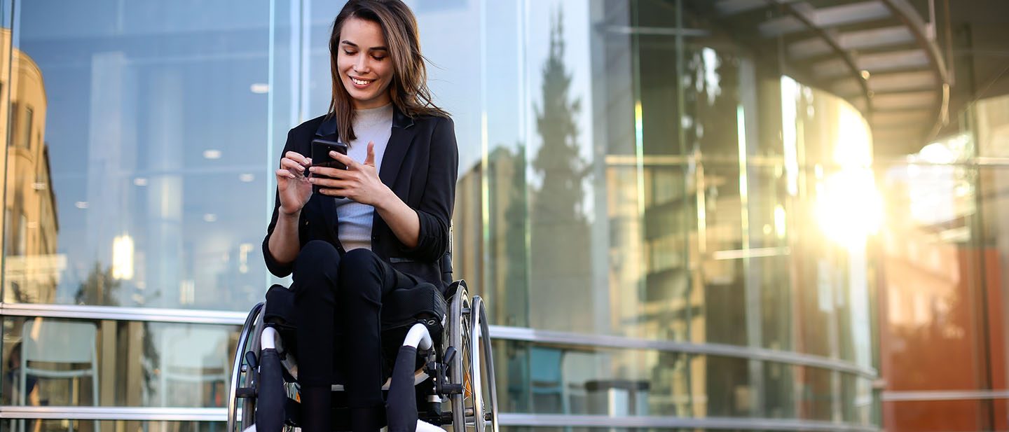 Woman in a wheelchair in front of the airport building looks at her cell phone.