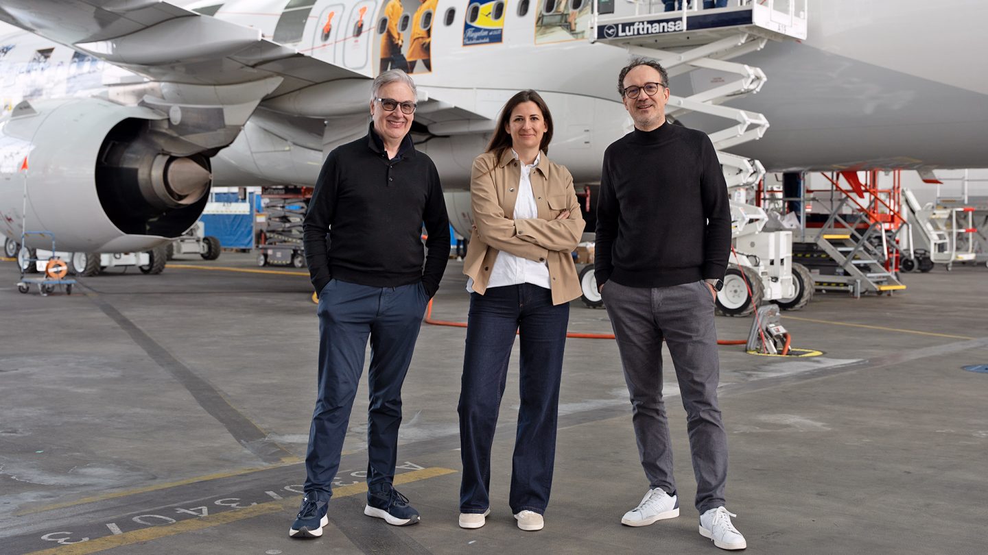 Three people are standing in front of a Lufthansa aircraft in a maintenance hangar, with work platforms in the background.