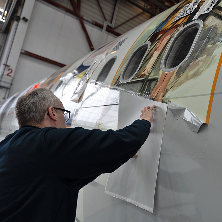 Technician applies graphic film to aircraft fuselage in a maintenance hangar.