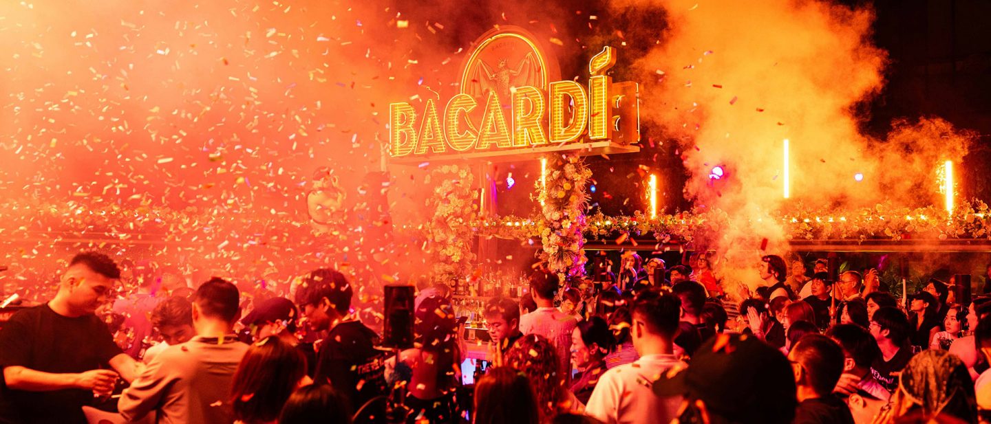 Crowd celebrating in front of a lit Bacardi bar as confetti, smoke, and orange light fill the scene.