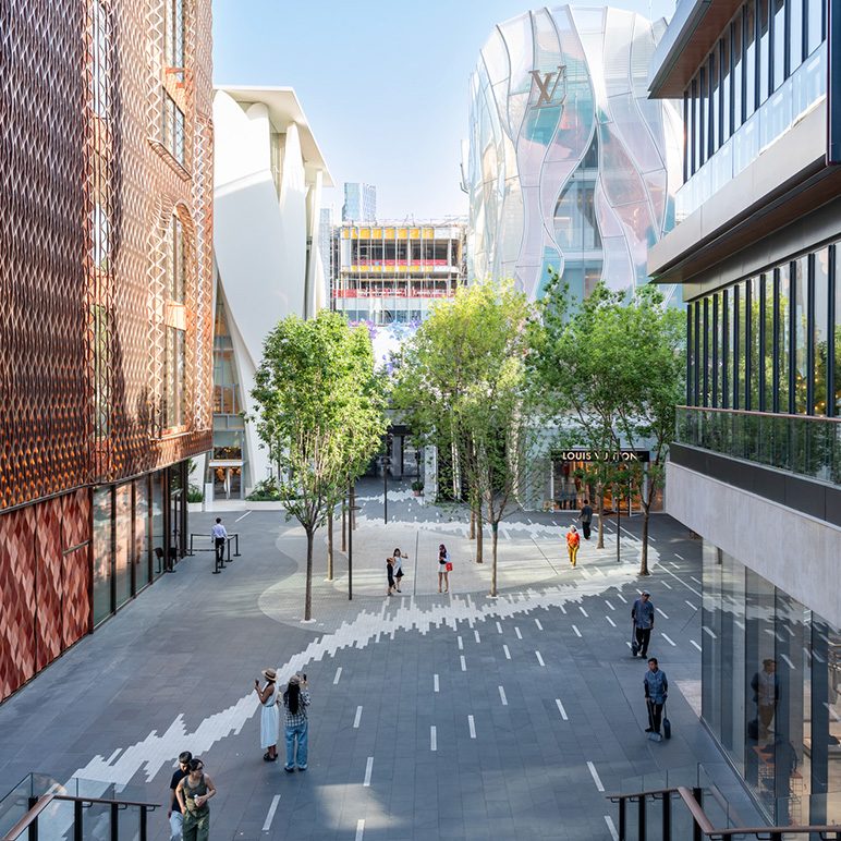 Open-air shopping plaza with trees, modern buildings, and a Louis Vuitton storefront.