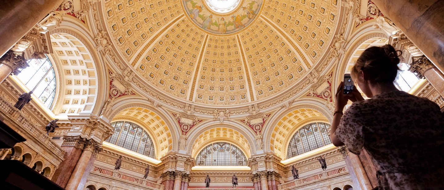 Photo of a woman capturing the ornate ceiling of a historic building in Washington, D.C., on her camera.