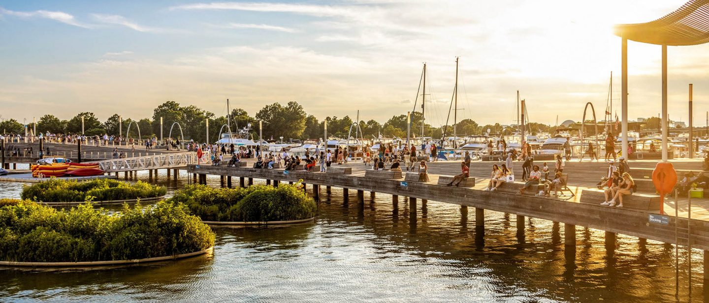 Picture of a bustling marina in Washington, D.C., with people enjoying sunset, featuring boats and a boardwalk.