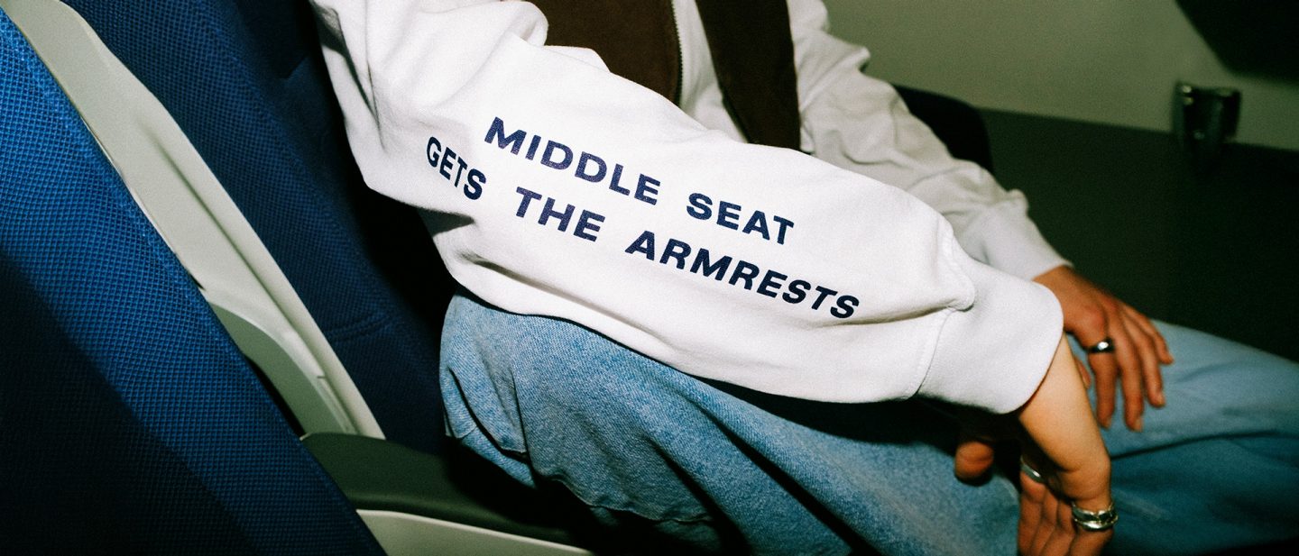 Person sitting in an airplane seat; sleeve text reads 'Middle seat gets the armrests.'
