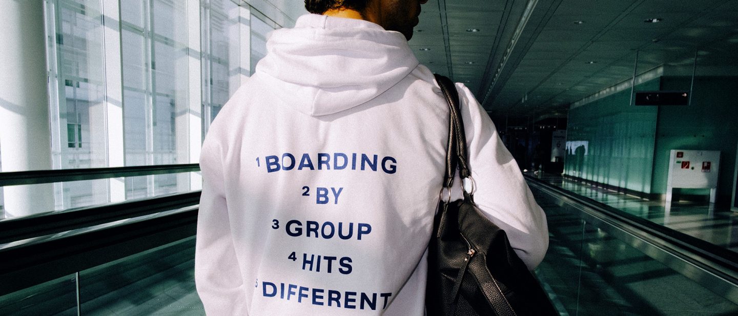 Back view of a person in an airport corridor wearing a hoodie with the text 'Boarding by group hits different' on the back.