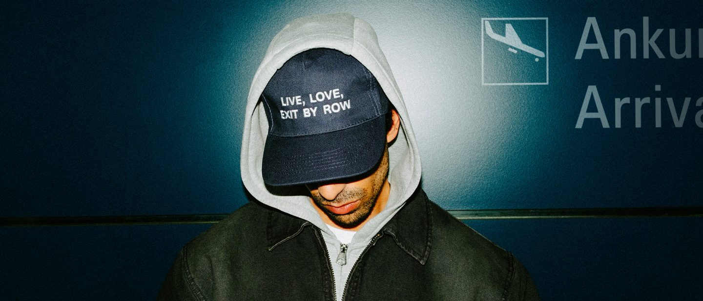 Person wearing a hoodie and cap in front of an airport wall; cap text reads “Live, love, exit by row.”