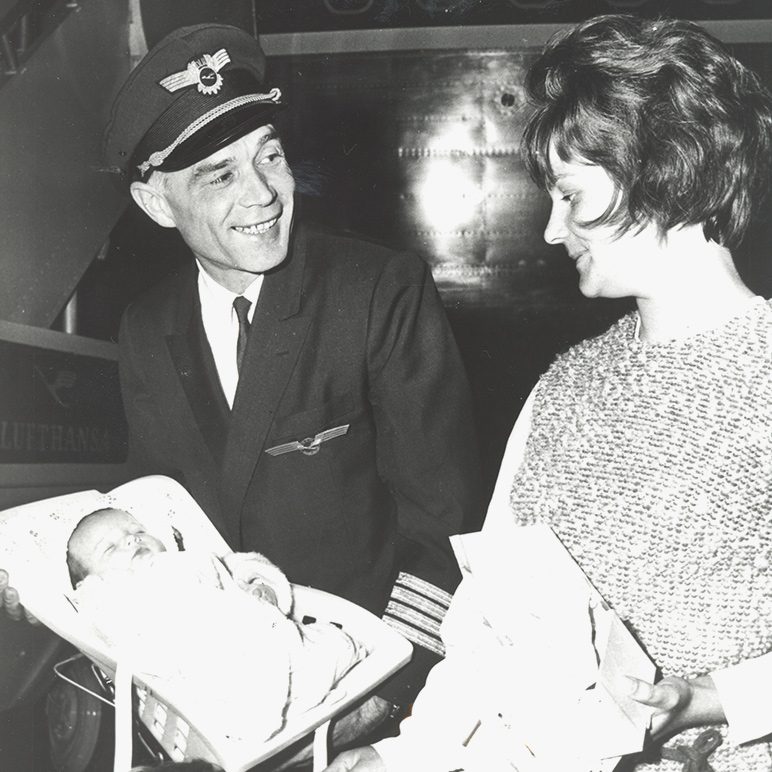 Black and white photo of a Lufthansa pilot in uniform, smiling while holding a newborn baby in a carrier