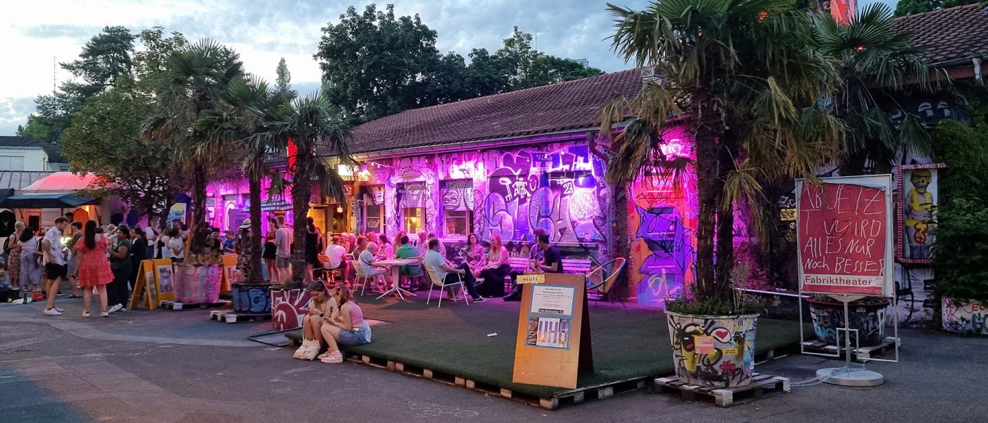 People at a vibrant street cafe with neon-lit graffiti art on the Rote Fabrik areal.