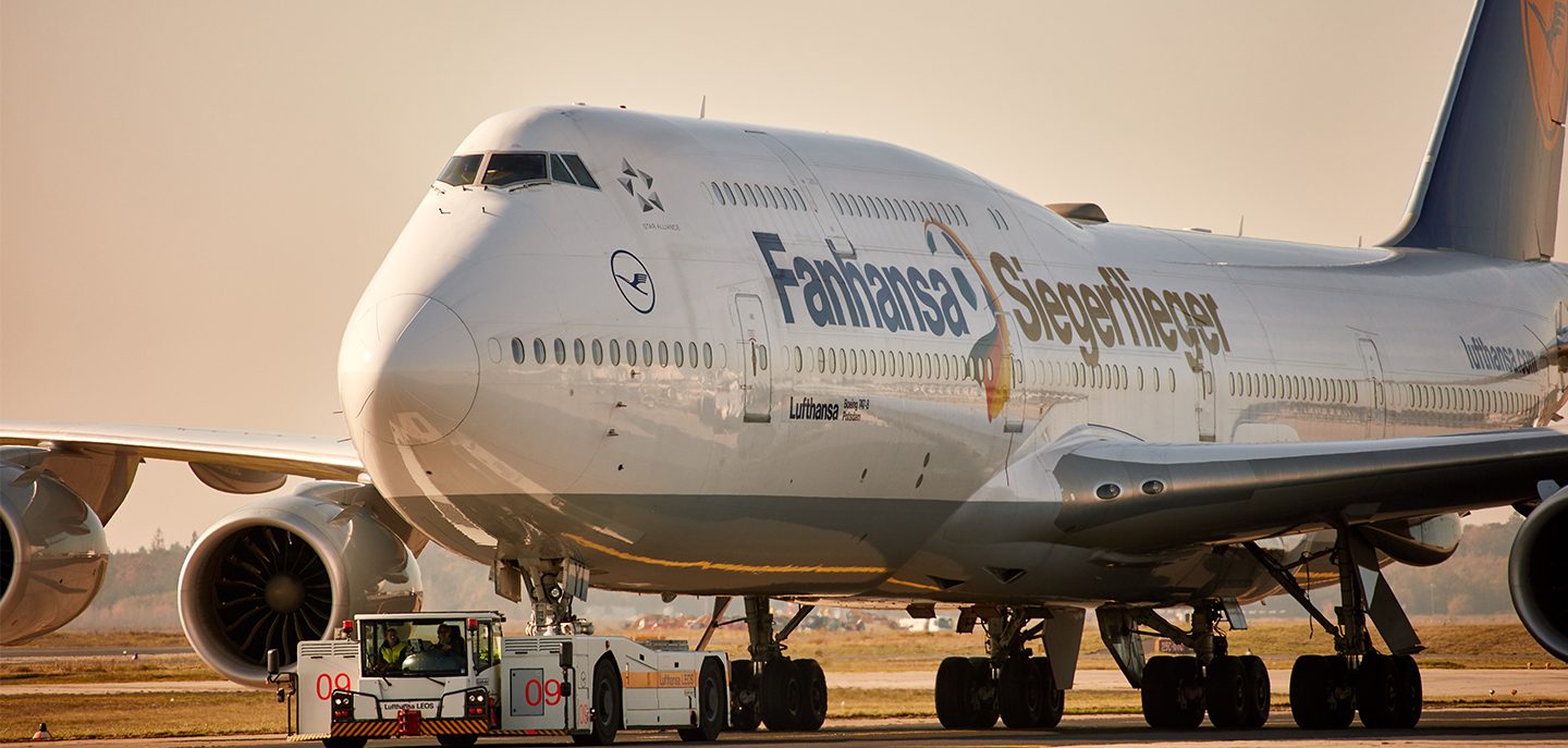 An airplane is standing on the runway and is being pulled by a vehicle. It bears the inscription Fanhansa Siegerflieger.