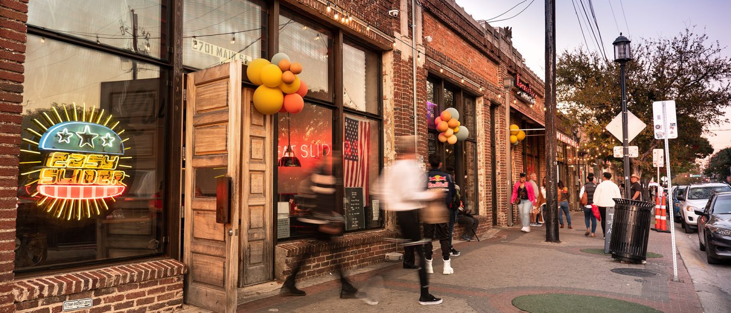 Busy street with a burger restaurant, neon sign, and pedestrians in front of brick buildings