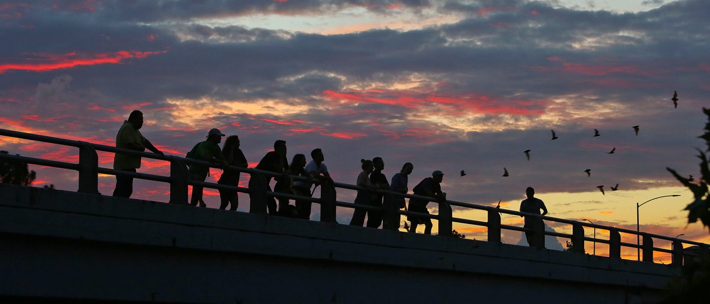 People stand silhouetted on a bridge against a colorful sunset sky
