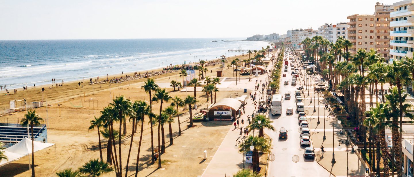 Busy beach promenade in Larnaca with palm trees, sandy beach, and hotels by the blue sea