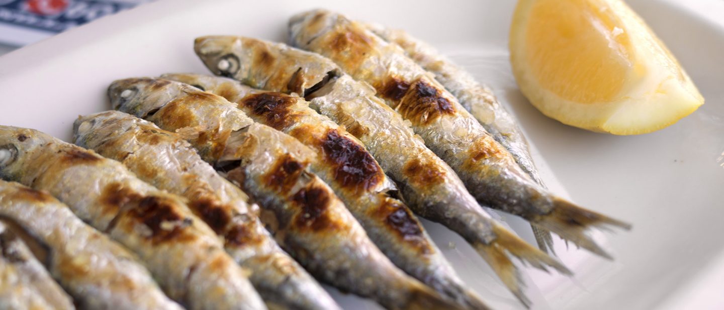 Freshly grilled sardines served on a white plate with a wedge of lemon