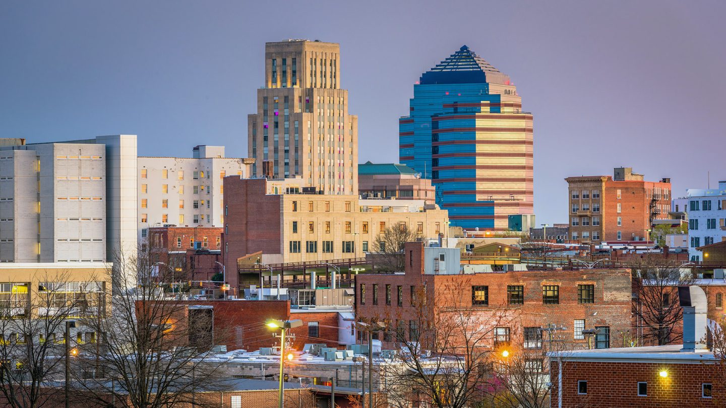 City skyline view of downtown buildings at dusk with clear sky and streetlights beginning to glow.