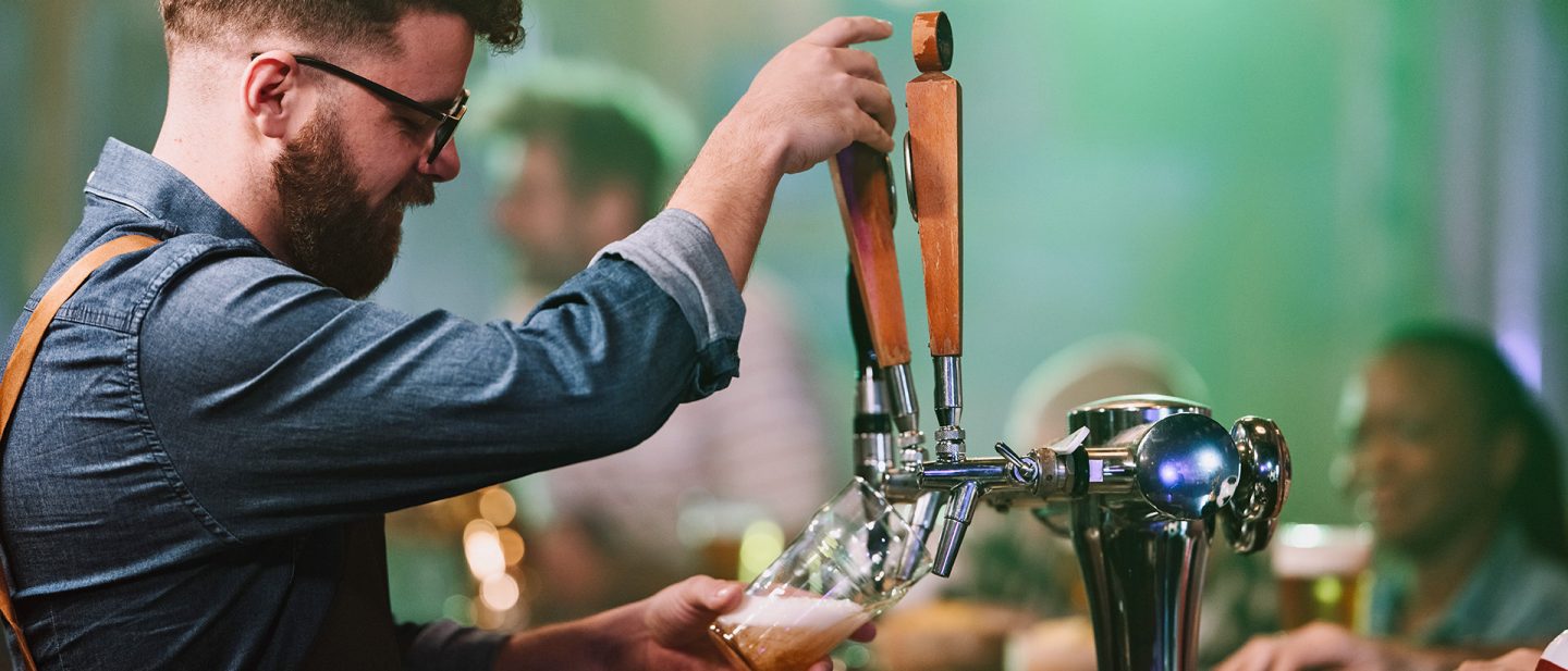 Man with glasses pouring draft beer from tap in a bar with patrons in the background.
