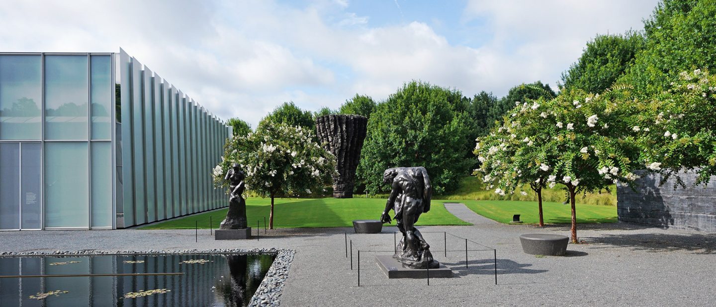 Outdoor sculpture garden with bronze statues, a modern glass building, flowering trees, and a reflective water feature under a blue sky.
