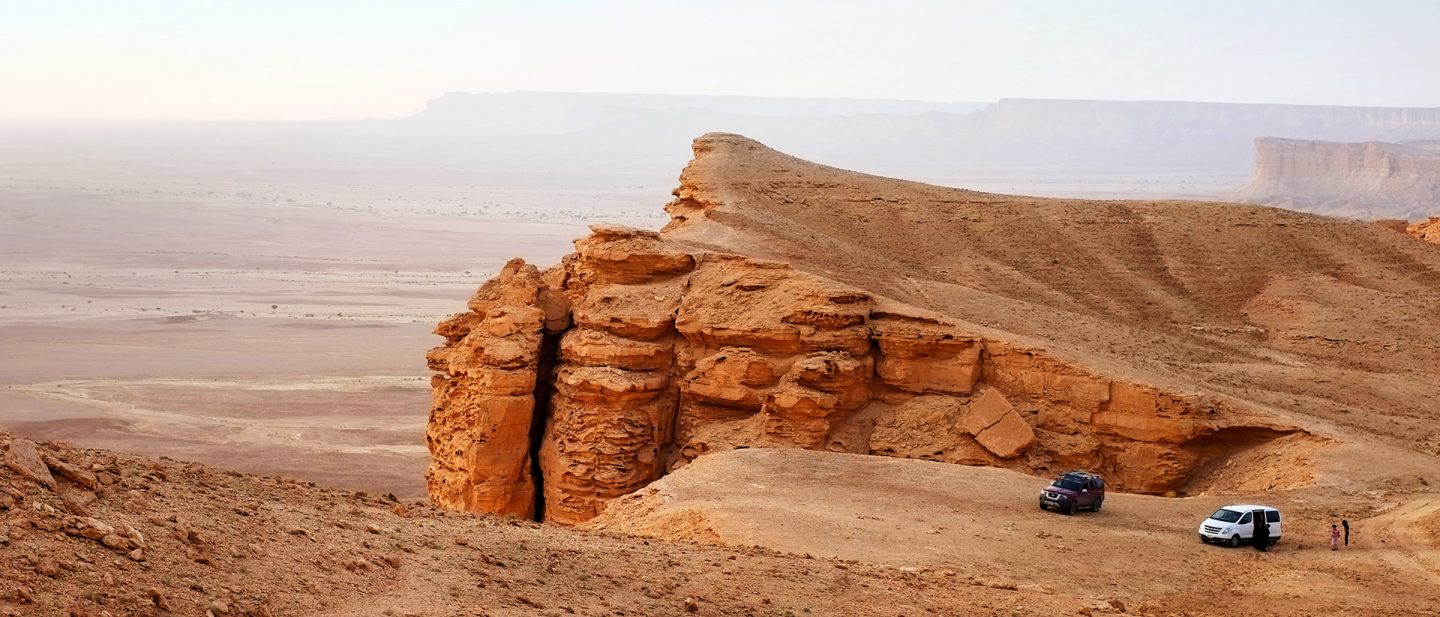 Off-road vehicles parked by a striking rock formation in a vast desert landscape under a hazy sky