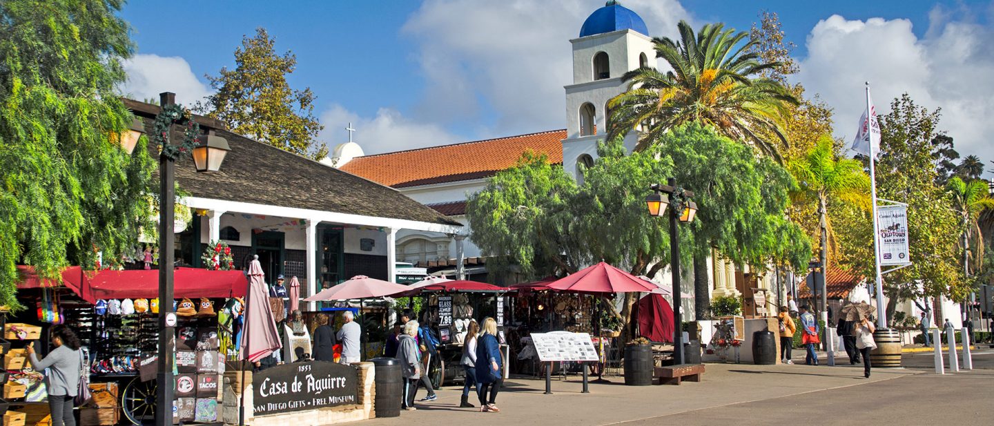 A bustling plaza in the Old Town San Diego State Historic Park featuring market stalls and a white church under palm trees