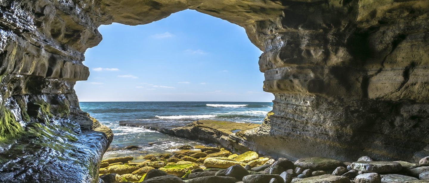 A view from inside a rocky sea cave looking out at the blue ocean and the coastline of La Jolla in San Diego