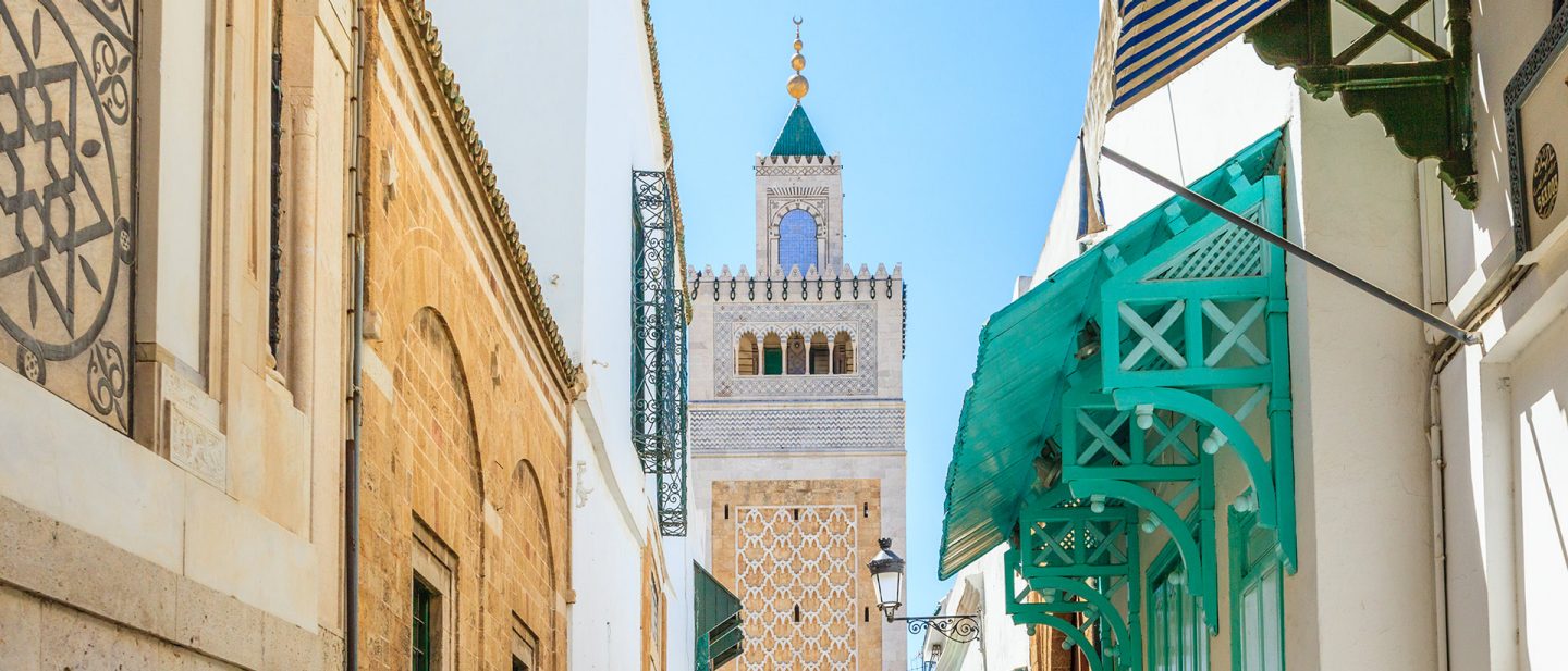 Straßenansicht traditioneller tunesischer Architektur mit einem Moscheeturm und bunten Holzbalkonen unter einem klaren blauen Himmel.