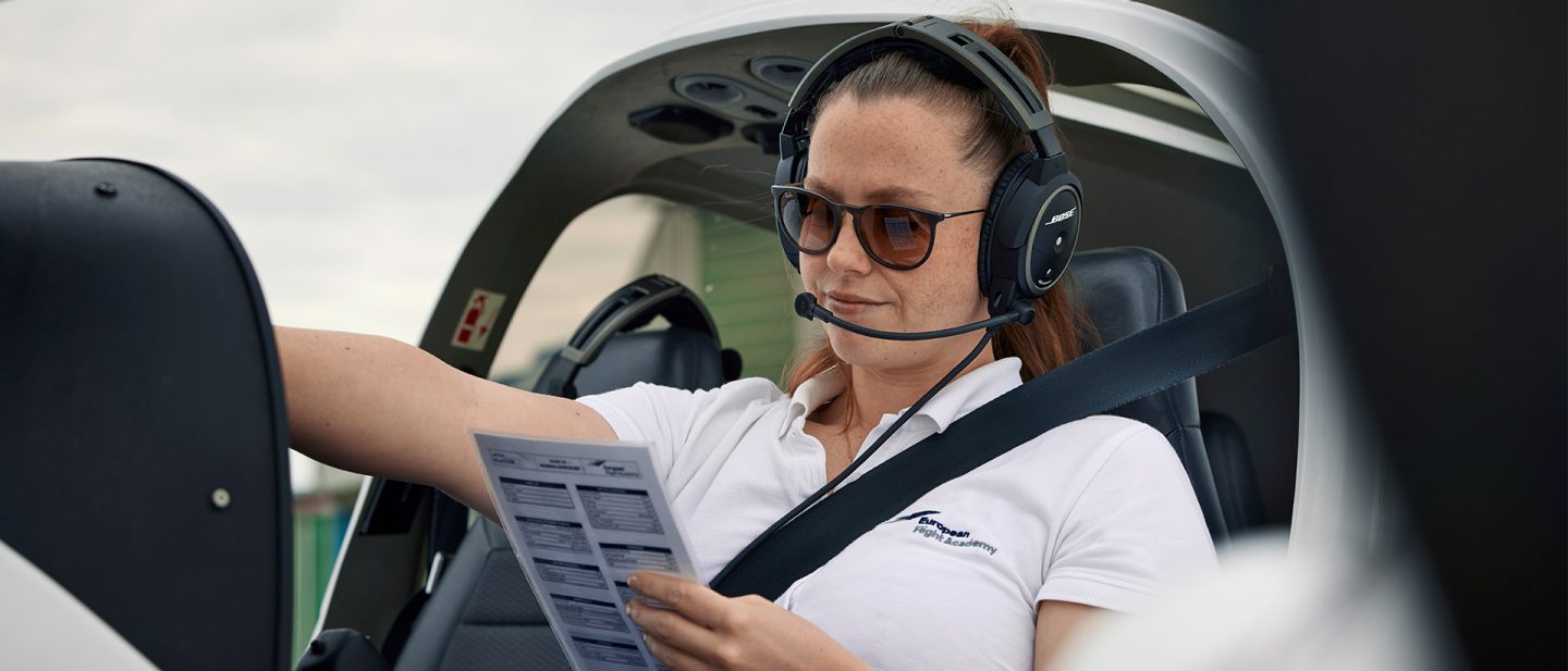 A female pilot sits strapped into the cockpit of a small aircraft, wearing a headset and sunglasses while checking a pre-flight checklist.